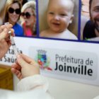 Vacinadora prepara dose para aplicação, ao fundo um cartaz com rosto de pessoas