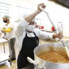 Mulher está na cozinha, mexendo uma panela e prepara refeição que será servida no Restaurante Popular
