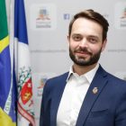 Jovita pousa para foto em frente as bandeiras e banner com brasão do município de Joinville