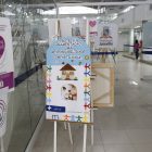 Cartaz tem o desenho em feltro mostrando uma casa e embaixo a foto de uma profissional da saúde auxiliando uma mãe a amamentar