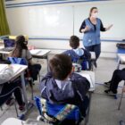 Professora em pé, em sala de aula, ensina alunos que estão em carteiras