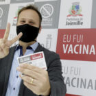 Prefeito de Joinville mostra carteira de vacinação em frente a banner logo após receber segunda dose