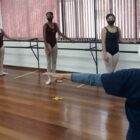 Professora dá aula de ballet para alunas apoiadas em uma barra