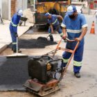Homens operam máquinas para reparar o pavimento de uma rua sinalizada com cones
