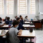 Professora em pé, na frente da sala de aula e alunos acompanhando a aula