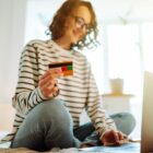 mulher jovem está sentada na cama e utiliza notebook enquanto segura um cartão de crédito