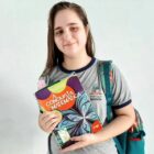 Aluna segura livro, tem uma mochila nas costas e posa para foto