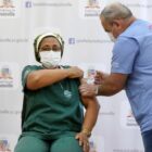 A enfermeira Rosangela sentada, de uniforme, recebe vacina de um profissional