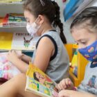 Duas meninas sentadas, com máscaras, leem livros