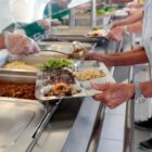 Homem segura bandeja e serve comida em um prato com macarrão, arroz, feijão e salada