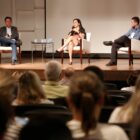 Três pessoas sentadas em cadeiras, em um palco, palestram para plateia