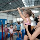 Professora segura aluna em barra de ginástica e outras alunas observam