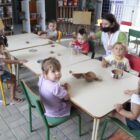 Seis crianças sentadas em roda, em cadeiras, fazem refeições enquanto são observadas por professoras