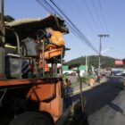Homens trabalham colocando asfalto em rua enquanto trabalhador dirige máquina para auxiliar o serviço