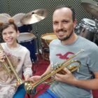 Aluna segura trompete sentada ao lado de professor que segura também um instrumento musical