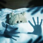 imagem de um urso de pelúcia na cama com olhos arregalados e sombras de mãos em cima