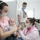 Mãe segura bebê no colo enquanto enfermeira aplica vacina e o pai da criança observa.