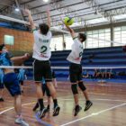 Meninos jogam volei em quadra, dois deles fazem bloqueio perto da rede