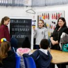 Alunos e professora, em sala de aula, apresentam trabalho para outros alunos sentados