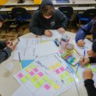Alunos sentados, escrevem e fazem atividades em uma mesa redonda