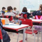 alunos em sala de aula, sentados em cadeiras, na frente de carteiras