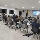 Pessoas sentadas em cadeiras, em uma sala, ouvem técnico