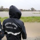 Dois agentes da Defesa Civil de casaco com capuz andam em rua alagada pela chuva