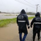 Dois agentes da Defesa Civil de casaco com capuz andam em rua alagada pela chuva