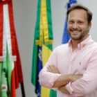 Prefeito Adriano Silva, de braços cruzados, na frente de bandeiras, olha para câmera