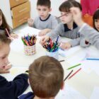 Alunos de educação infantil com uniforme, brincam ao redor de mesa com lápis de cor e papéis