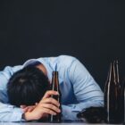 Homem com a cabeça abaixada em uma mesa segura uma garrafa e há outras ao lado