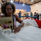 Costureira trabalha em máquina de costura e atrás dela integrantes da escola ensaiam