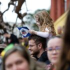 Menina carregada pelo pai segura bandeira de Joinville enquanto público acompanha desfile