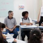 Alunos sentados em frente de carteiras, em roda, prestam atenção em professora que está em pé