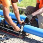 Homens, uniformizados e com equipamentos de segurança, trabalham em tubulação