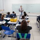 Professora em pé, em sala de aula, ensina alunos que estão em carteiras