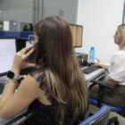 Duas mulheres trabalham sentadas em frente a computadores, uma delas segura um telefone celular