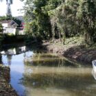 Área com um rio, vegetação ao lado, um barquinho na água e uma casa ao fundo