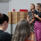 Duas mulheres em pé, uma fala ao microfone, olhando para plateia
