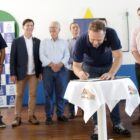 Prefeito assina documento em mesa e é observado por outras autoridades