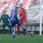 Jogador chuta bola no gol, com zagueiro e goleiro na frente