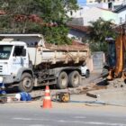 Dois homens, com capacetes e uniformes, trabalham em rua, ao lado de caminhão e máquinas