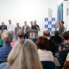 Prefeito discursa em púlpito para plateia sentada e autoridades em pé