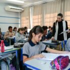 Em sala de aula, professora está entre carteiras e alunos sentados em prova