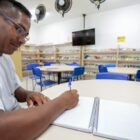 Homem moreno com óculos escreve em caderno e sorri em sala de aula