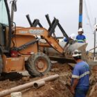 Homens, com uniformes e capacetes, trabalham ao lado de retroescavadeira