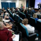 Profissionais da educação sentados em plateia observam e escutam secretário em apresentação do programa