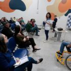 Pessoas reunidas em sala onde palestrante está em pé ao lado de um cadeirante