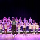 Alunos se apresentam em palco, lado a lado, cantando, com professora a frente
