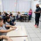 Agentes da Defesa Civil em pé na frente de sala de aula falam para alunos sentados em carteiras
